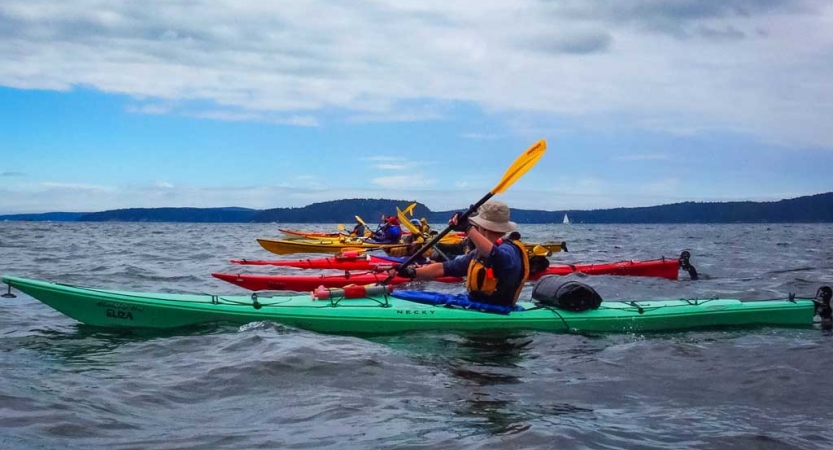 a line of colorful kayaks are paddled by outward bound students on a calm body of water. There appear to be mountains in the background.